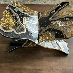 May include: A decorative, open book-shaped stand with a black and gold design. The stand features a black base and a top with a glittery black and gold pattern, accented with silver and small white floral details. The stand is made of resin.