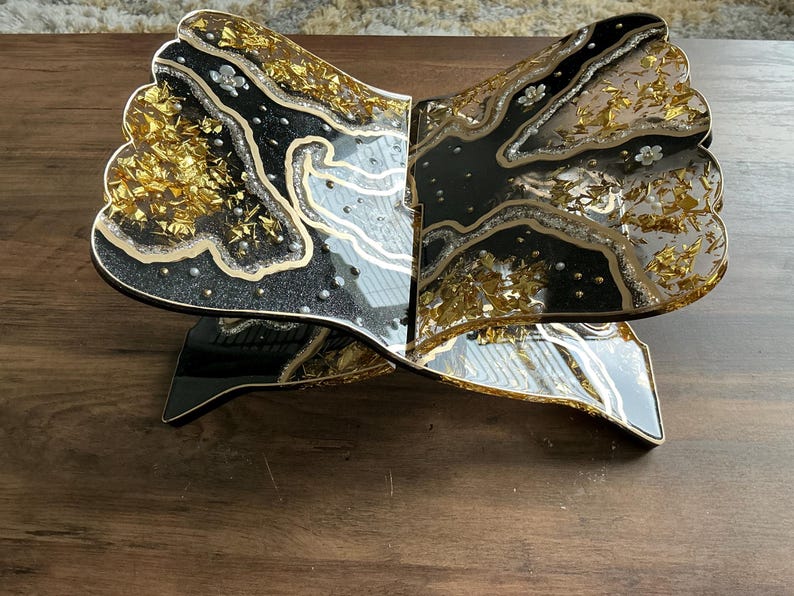 May include: A decorative, open book-shaped stand with a black and gold design. The stand features a black base and a top with a glittery black and gold pattern, accented with silver and small white floral details. The stand is made of resin.