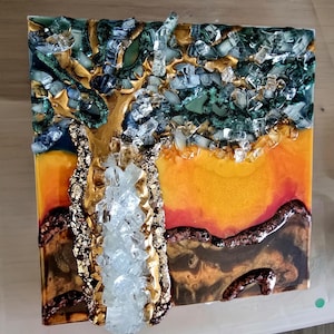K&ouml;nnte beinhalten: Quadratisches Kunstwerk aus Kunstharz mit einem Baum mit goldenem Stamm und &Auml;sten, verziert mit klaren und blauen Glasst&uuml;cken. Der Hintergrund wechselt von Orange zu Gelb, mit braunen und goldenen Akzenten am unteren Rand.
