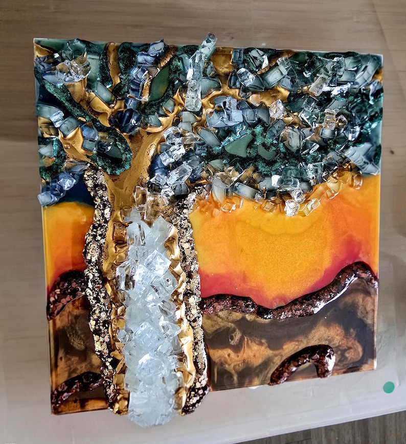 K&ouml;nnte beinhalten: Quadratisches Kunstwerk aus Kunstharz mit einem Baum mit goldenem Stamm und &Auml;sten, verziert mit klaren und blauen Glasst&uuml;cken. Der Hintergrund wechselt von Orange zu Gelb, mit braunen und goldenen Akzenten am unteren Rand.