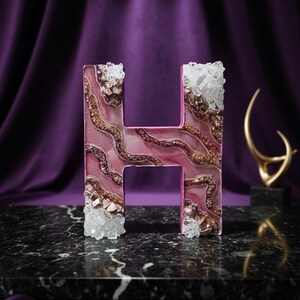 May include: A decorative letter "H" in pink resin, adorned with gold and crystal accents. The letter is set against a purple backdrop and rests on a black marble surface, creating a luxurious aesthetic.