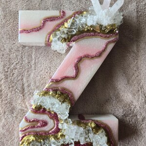 May include: A decorative letter "Z" in a light pink and white color scheme, embellished with gold and pink glitter accents. Clear crystal and white crystal clusters are embedded along the edges, creating a unique, textured design.