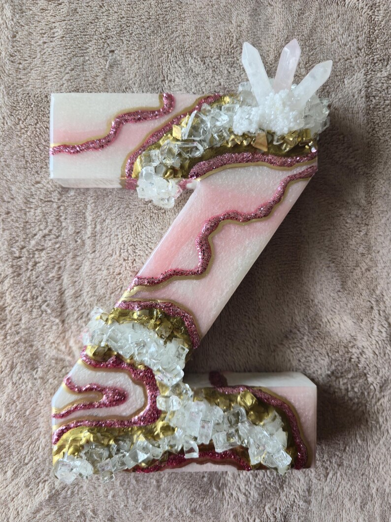 May include: A decorative letter "Z" in a light pink and white color scheme, embellished with gold and pink glitter accents. Clear crystal and white crystal clusters are embedded along the edges, creating a unique, textured design.