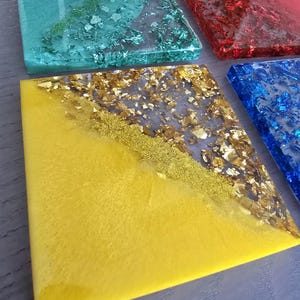Pode incluir: Quatro bases quadradas de resina nas cores verde-azulado, vermelho, azul e amarelo. Cada base apresenta flocos met&aacute;licos e glitter embutidos, criando um efeito brilhante. As bases est&atilde;o dispostas sobre uma superf&iacute;cie cinza.