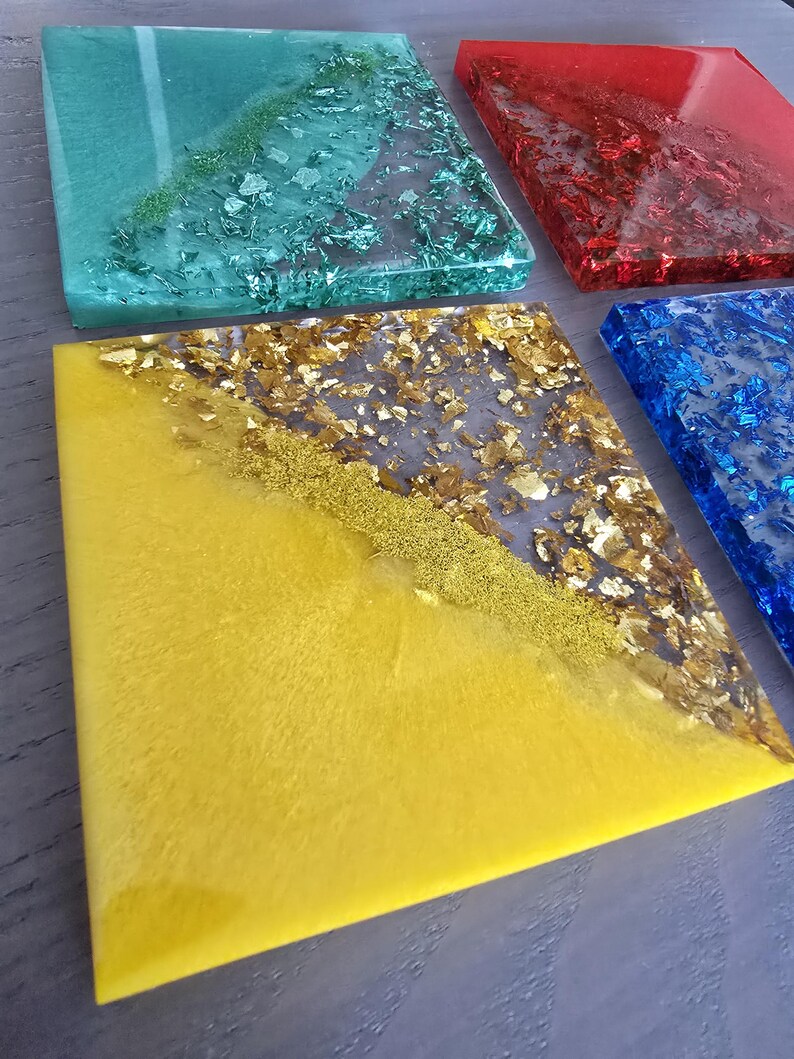 Pode incluir: Quatro bases quadradas de resina nas cores verde-azulado, vermelho, azul e amarelo. Cada base apresenta flocos met&aacute;licos e glitter embutidos, criando um efeito brilhante. As bases est&atilde;o dispostas sobre uma superf&iacute;cie cinza.