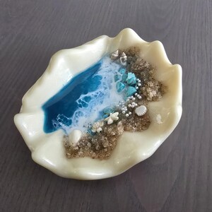 May include: An off-white seashell-shaped dish with a miniature beach scene. The scene features a blue resin ocean, sand, turquoise stones, tiny shells, pearls, and a starfish. The dish is on a grey surface.