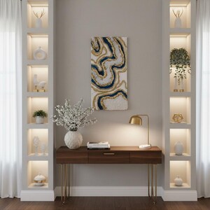 Op de afbeelding: Een modern interieur met een donkerhouten bureau met gouden poten, een gouden bureaulamp en een witte vaas met bloemen. Een abstract kunstwerk hangt boven het bureau. Ingebouwde planken tonen decoratieve items.