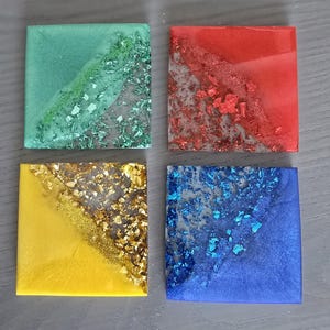 Pode incluir: Quatro bases quadradas nas cores verde, vermelho, amarelo e azul. Cada base tem um design diagonal com glitter e flocos met&aacute;licos. As bases s&atilde;o feitas de resina e est&atilde;o sobre uma superf&iacute;cie cinza.