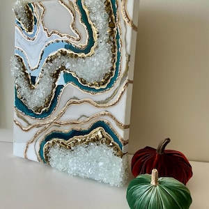 May include: A white canvas art piece with a geode design featuring teal, gold, and light blue accents. The artwork has a textured, crystal-like appearance. Two velvet pumpkins, one red and one green, are placed in front of the artwork.