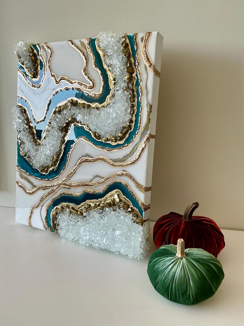 May include: A white canvas art piece with a geode design featuring teal, gold, and light blue accents. The artwork has a textured, crystal-like appearance. Two velvet pumpkins, one red and one green, are placed in front of the artwork.