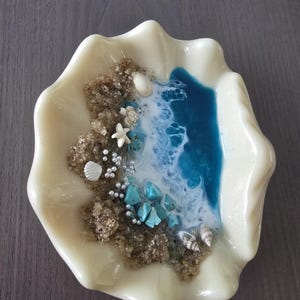 May include: An off-white, seashell-shaped dish featuring a miniature beach scene. It includes sand, blue resin representing water, small seashells, turquoise stones, and tiny white pearls. A unique coastal-themed decorative item for the home.