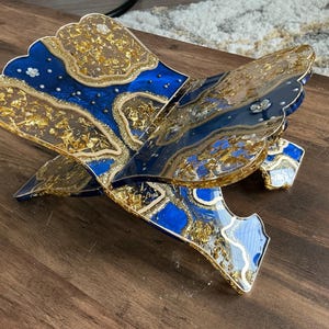 May include: A decorative, openwork book stand with a blue and gold design. The stand features a layered design with gold leaf accents and small white floral embellishments. The stand is made of resin and is displayed on a wooden surface.