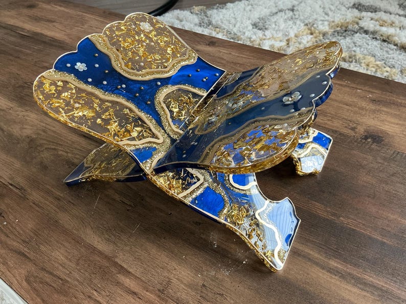 May include: A decorative, openwork book stand with a blue and gold design. The stand features a layered design with gold leaf accents and small white floral embellishments. The stand is made of resin and is displayed on a wooden surface.