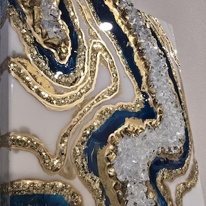 May include: Abstract resin art piece with a geode design. The artwork features layers of white, deep blue, and gold glitter resin, with clear crystal accents. The piece has a glossy finish and a textured appearance, creating a luxurious aesthetic.