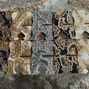May include: A collection of decorative phone stands in various colors and designs. Each stand is shaped like the letter "A" and features a unique pattern with gold leaf accents. Colors include blue, white, silver, black, and gold.