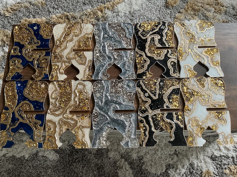 May include: A collection of decorative phone stands in various colors and designs. Each stand is shaped like the letter "A" and features a unique pattern with gold leaf accents. Colors include blue, white, silver, black, and gold.