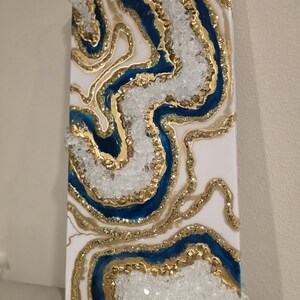 May include: Abstract wall art featuring a geode design. The artwork showcases a white background with flowing patterns of gold glitter, deep blue resin, and clear, crystalline accents. The piece is rectangular and has a three-dimensional appearance.
