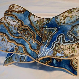 May include: A decorative wooden book stand with a unique design. The stand features a blue and white resin design with gold leaf accents. The edges are trimmed with gold and brown. The stand is designed to hold a book open.