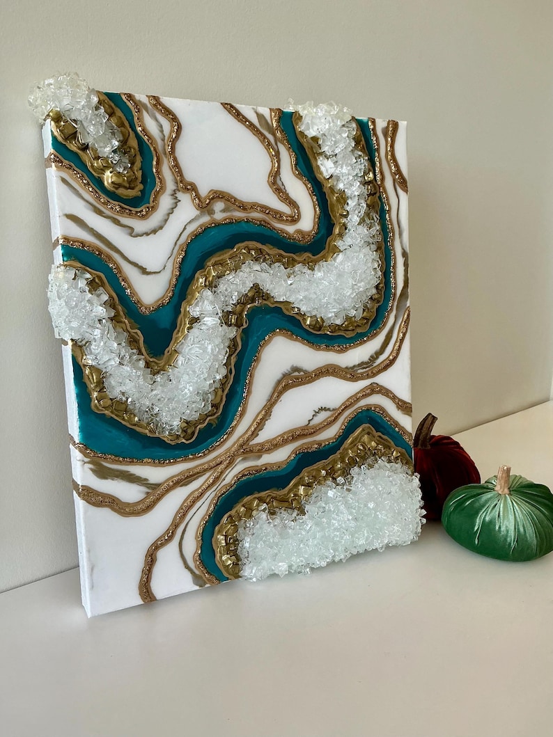 May include: Abstract canvas art with a geode design. The artwork features white, teal, and gold resin layers, with clear crystal accents. The canvas is rectangular and has a textured appearance, creating a unique decorative piece.