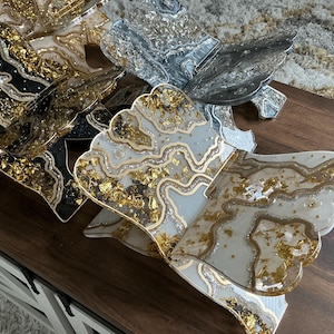 May include: A collection of decorative resin trays with a unique, layered design. The trays feature a variety of colors, including black, silver, and white, with gold leaf accents. Each tray has a scalloped edge and a glossy finish, creating an elegant aesthetic.