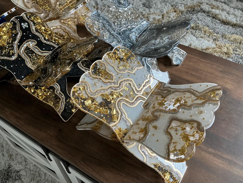 May include: A collection of decorative resin trays with a unique, layered design. The trays feature a variety of colors, including black, silver, and white, with gold leaf accents. Each tray has a scalloped edge and a glossy finish, creating an elegant aesthetic.