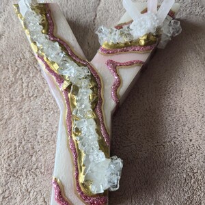 May include: A decorative letter "Y" in a geode style. The letter is white with pink glitter accents and gold trim. Clear crystals and quartz clusters are embedded throughout the design, creating a unique, eye-catching piece.