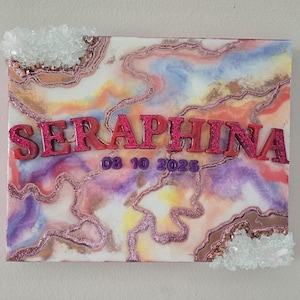 Puede incluir: Un lienzo rectangular con un dise&ntilde;o abstracto colorido en tonos rosa, morado, amarillo y azul. El nombre "SERAPHINA" se muestra en letras rosas brillantes, con la fecha "08 10 2025" debajo. Adornos cristalinos transparentes adornan los bordes.