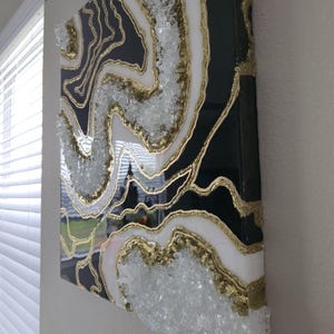 May include: Abstract geode-style wall art. The artwork features a black background with white and gold accents, and clear crystal embellishments. The piece is mounted on a rectangular canvas.