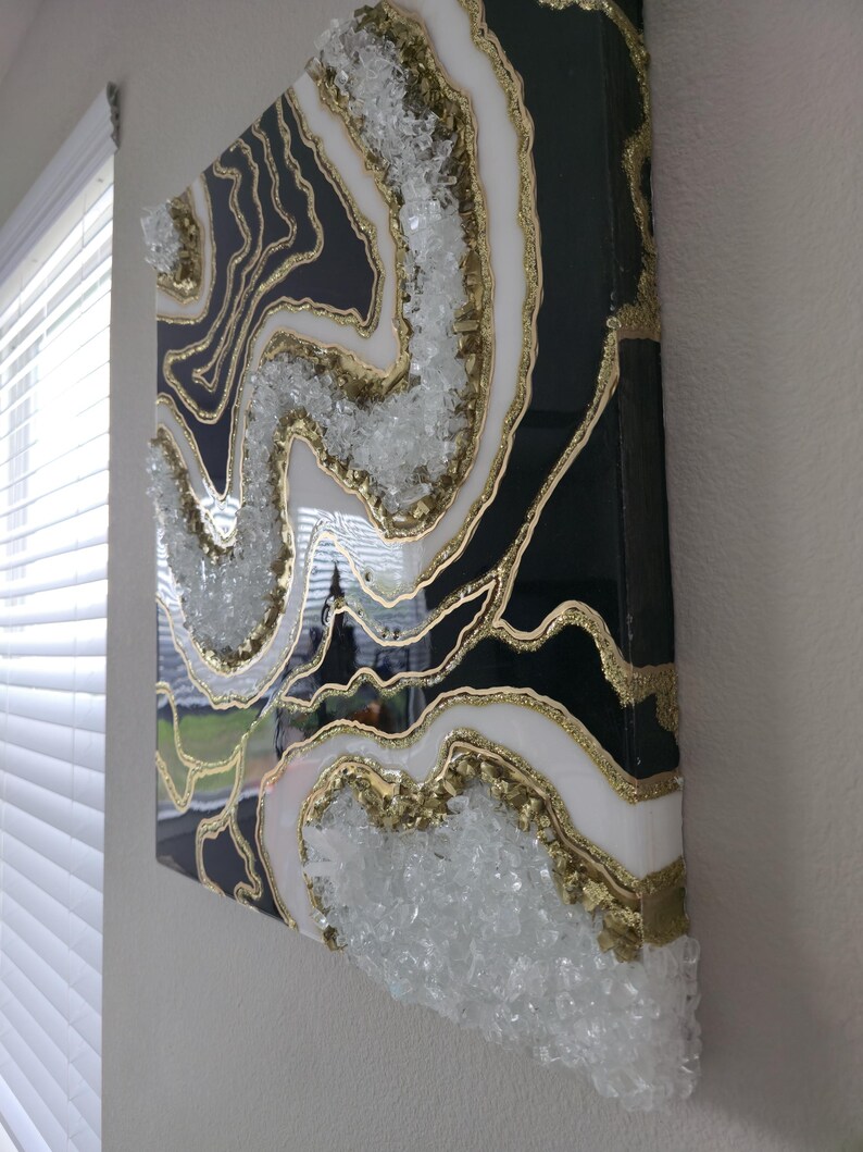 May include: Abstract geode-style wall art. The artwork features a black background with white and gold accents, and clear crystal embellishments. The piece is mounted on a rectangular canvas.