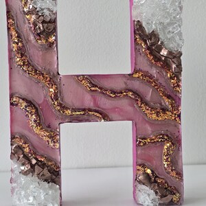 May include: A decorative letter "H" in a pink hue, adorned with gold glitter, brown metallic accents, and clear, crystalline embellishments. The letter has a textured, layered design, creating a unique, artistic appearance. The edges are a solid pink color.