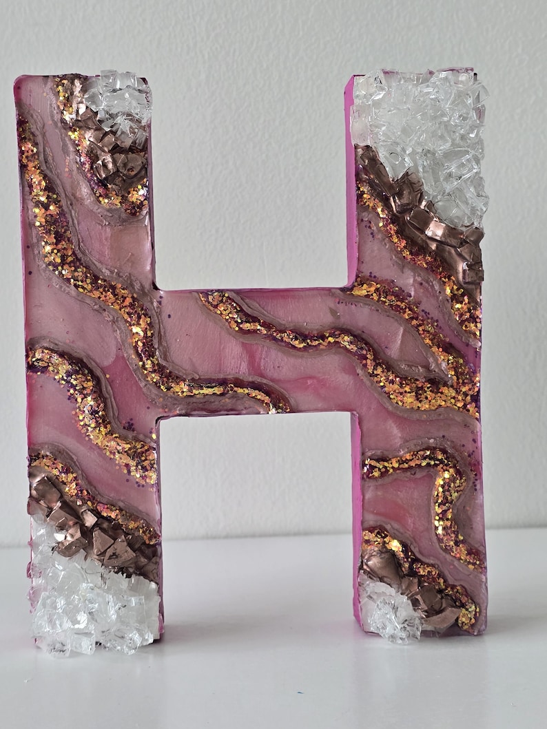 May include: A decorative letter "H" in a pink hue, adorned with gold glitter, brown metallic accents, and clear, crystalline embellishments. The letter has a textured, layered design, creating a unique, artistic appearance. The edges are a solid pink color.