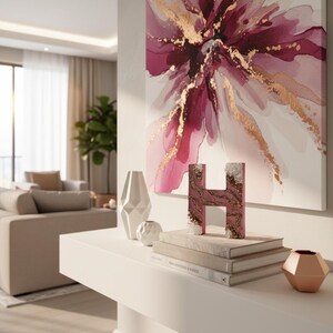 May include: A modern living room scene featuring a large abstract floral painting in shades of pink and gold. A white shelf displays decorative items, including a letter "H" sculpture, books, and geometric vases. The room has neutral tones and natural light.