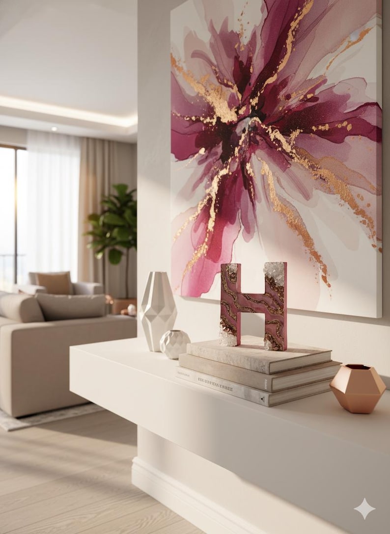 May include: A modern living room scene featuring a large abstract floral painting in shades of pink and gold. A white shelf displays decorative items, including a letter "H" sculpture, books, and geometric vases. The room has neutral tones and natural light.