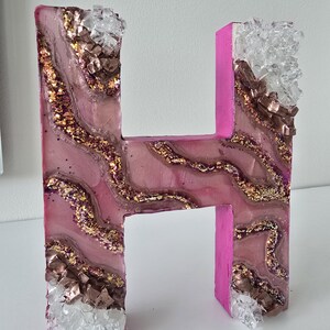 May include: A decorative letter "H" in pink, with a resin design featuring gold glitter and brown and clear crystal accents. The letter is standing upright, showcasing its unique, handcrafted appearance.