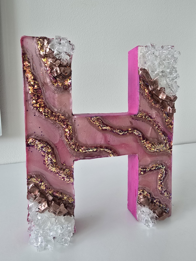 May include: A decorative letter "H" in pink, with a resin design featuring gold glitter and brown and clear crystal accents. The letter is standing upright, showcasing its unique, handcrafted appearance.