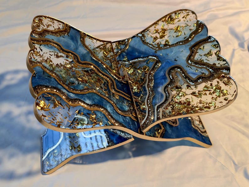 May include: A decorative wooden book stand with a unique design. The stand features a blue and white marbled pattern with gold leaf accents. The edges are outlined in gold. The stand is designed to hold a book open.