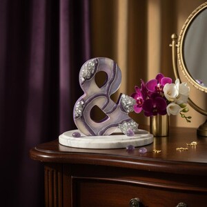 May include: A decorative ampersand sculpture in shades of purple and white, adorned with sparkling crystals. The sculpture sits on a marble base, displayed on a dark wooden table. A small vase of flowers and a vanity mirror are in the background.