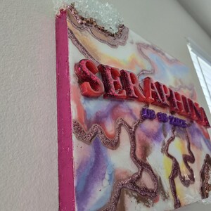 Puede incluir: Una pieza de arte mural decorativa con la palabra "SERAPHINA" en letras rojas en relieve. La obra presenta un dise&ntilde;o colorido y veteado con detalles de purpurina y un grupo de cristales transparentes en la parte superior. Los lados est&aacute;n pintados de rosa.