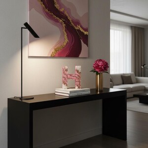 May include: A modern interior scene featuring a black console table with decorative items. A pink and gold abstract painting hangs above. On the table are a decorative letter "H", books, a gold vase with a pink flower, and a black lamp.