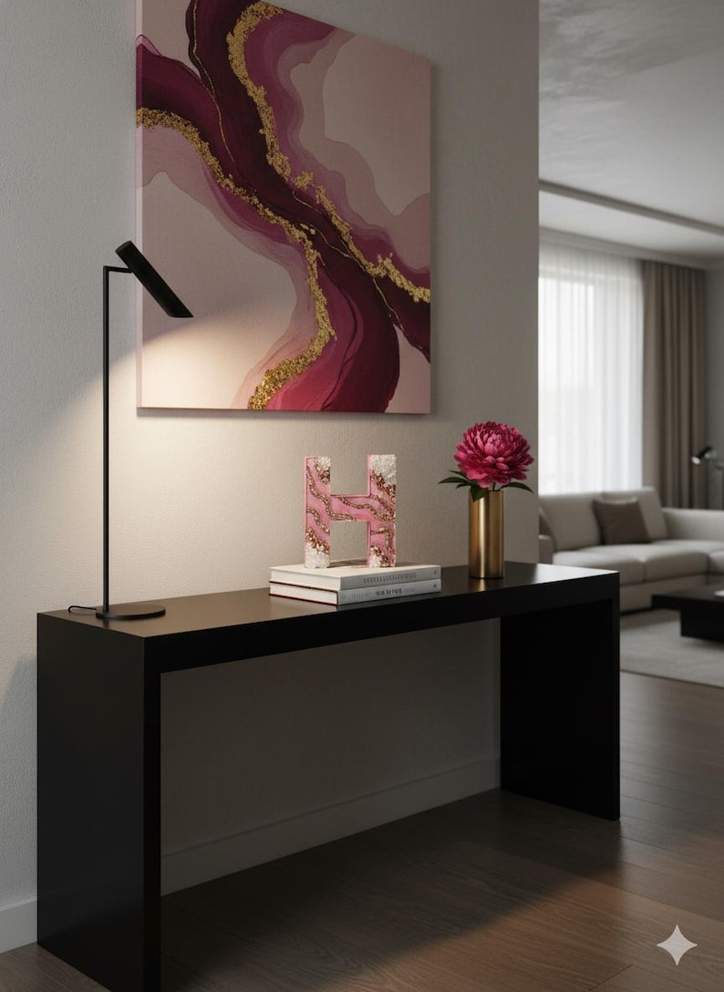 May include: A modern interior scene featuring a black console table with decorative items. A pink and gold abstract painting hangs above. On the table are a decorative letter "H", books, a gold vase with a pink flower, and a black lamp.