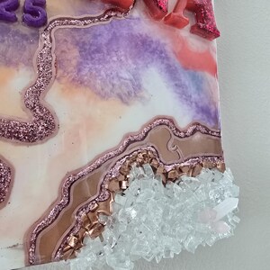 Puede incluir: Una obra de arte decorativa con el nombre "TINA" en letras de purpurina rosa y el n&uacute;mero "25" en morado. La pieza presenta un dise&ntilde;o en capas con tonos rosas, morados y blancos, realzado con purpurina y adornos de cristal transparente.