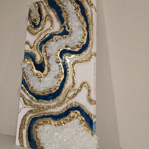 Op de afbeelding: Abstract kunstwerk van hars met een witte achtergrond, blauwe en gouden accenten. Het ontwerp bootst een geode na, met lagen hars en heldere, kristalachtige elementen. Het rechthoekige kunstwerk is op een wit oppervlak gemonteerd.