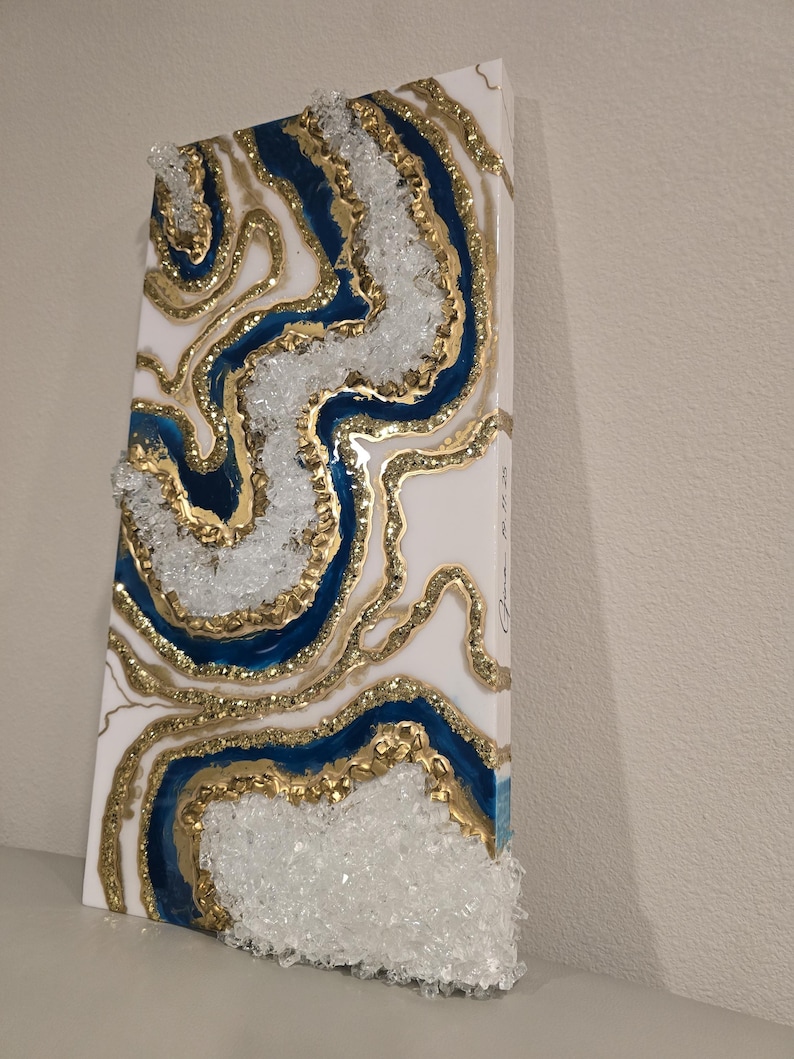 Op de afbeelding: Abstract kunstwerk van hars met een witte achtergrond, blauwe en gouden accenten. Het ontwerp bootst een geode na, met lagen hars en heldere, kristalachtige elementen. Het rechthoekige kunstwerk is op een wit oppervlak gemonteerd.