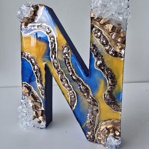 Puede incluir: Una letra "N" decorativa de madera con un dise&ntilde;o de resina azul, amarillo y dorado. La letra est&aacute; adornada con purpurina y detalles de cristal. Los bordes son de color azul oscuro.