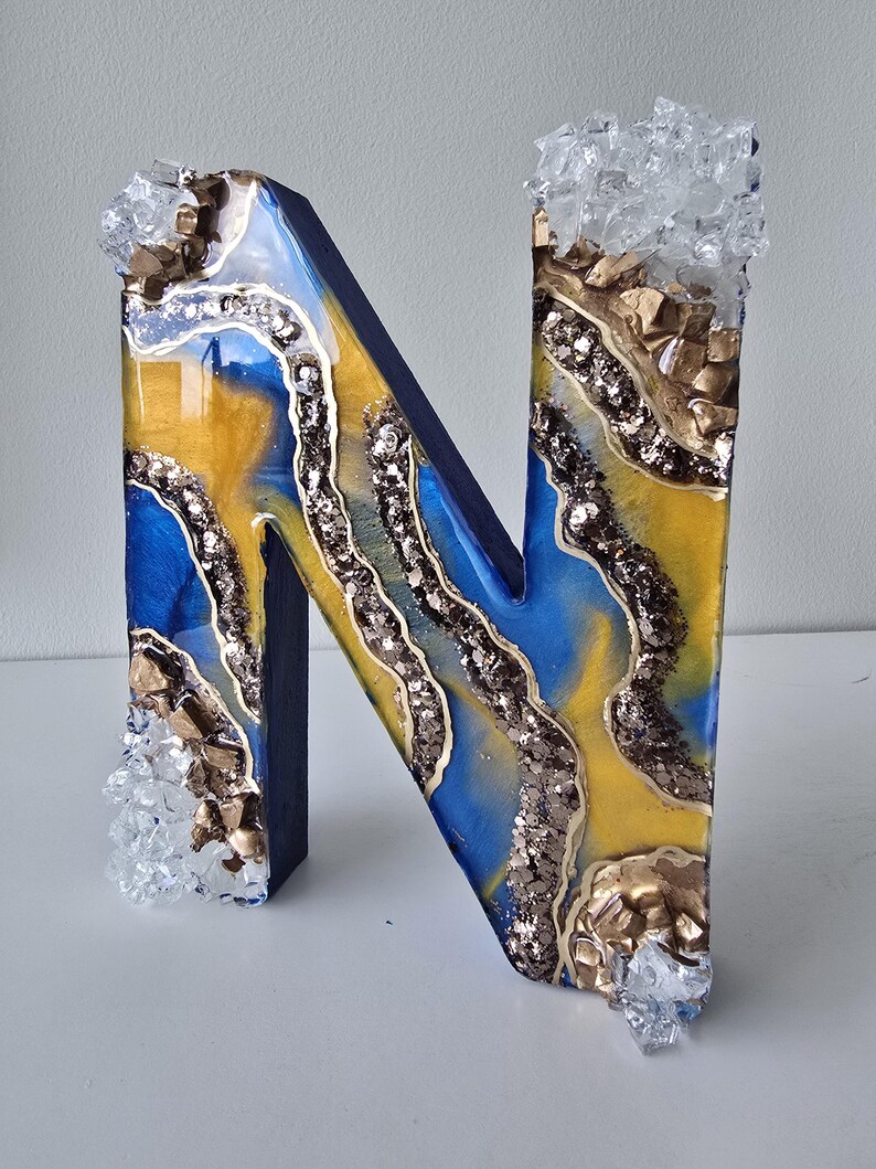 Puede incluir: Una letra "N" decorativa de madera con un dise&ntilde;o de resina azul, amarillo y dorado. La letra est&aacute; adornada con purpurina y detalles de cristal. Los bordes son de color azul oscuro.