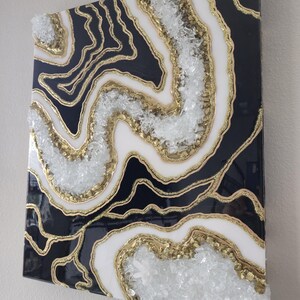 May include: Abstract wall art with a geode design. The artwork features a black, white, and gold colour scheme, with clear crystal accents. The design resembles natural stone formations, creating a unique decorative piece.