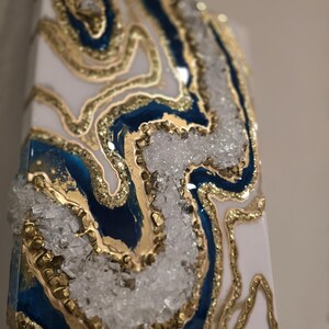 May include: Abstract art piece with a geode design. The artwork features a white base with blue and gold accents, and clear crystal-like embellishments. The piece has a rectangular shape.