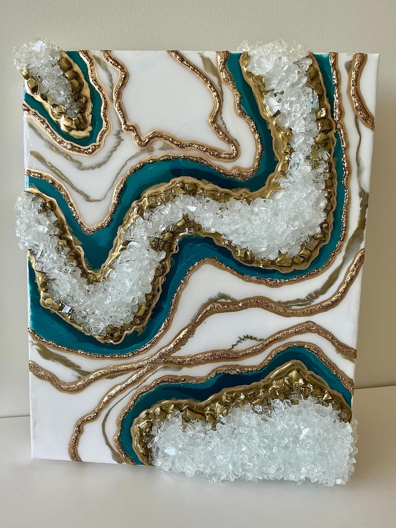 May include: Abstract art piece featuring a white background with teal and gold accents, resembling a geode. Clear, crystalline embellishments are applied to the surface, adding texture and dimension to the artwork. The piece is mounted on a canvas.