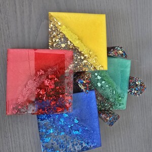 Pode incluir: Vista a&eacute;rea de cinco bases quadradas de resina nas cores vermelha, amarela, azul e verde. Cada base &eacute; incrustada com flocos met&aacute;licos. A base amarela tem uma faixa dourada. Aproximadamente 8 cm x 8 cm.