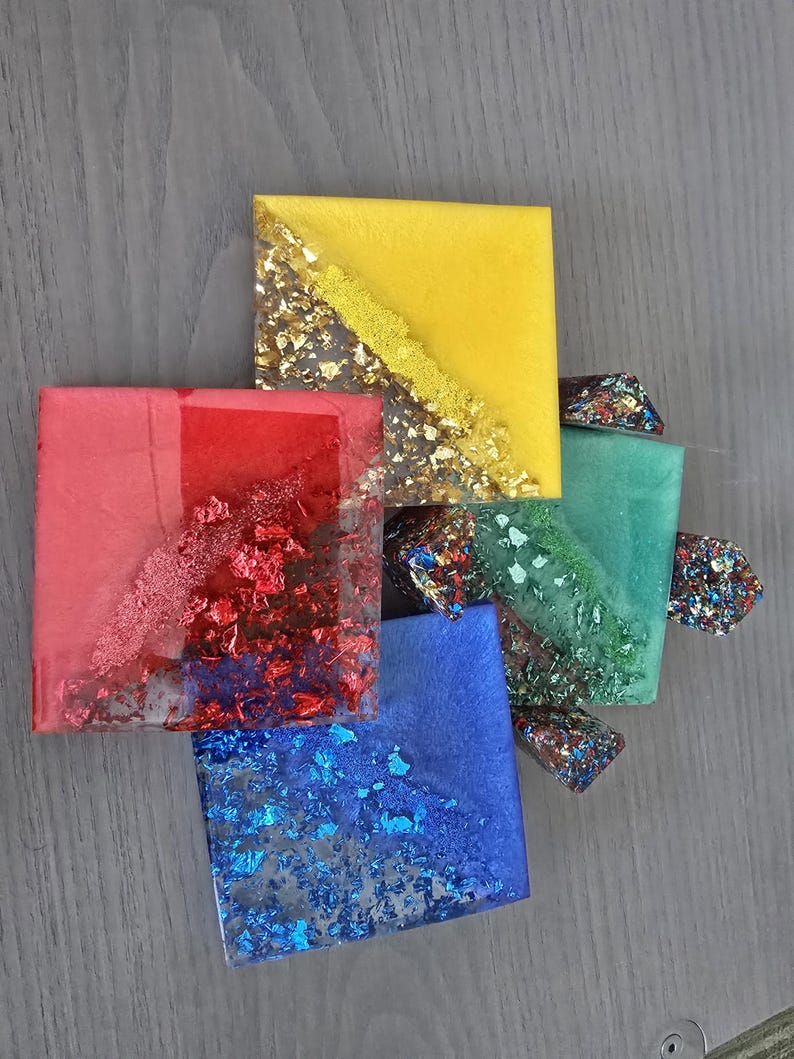 Pode incluir: Vista a&eacute;rea de cinco bases quadradas de resina nas cores vermelha, amarela, azul e verde. Cada base &eacute; incrustada com flocos met&aacute;licos. A base amarela tem uma faixa dourada. Aproximadamente 8 cm x 8 cm.